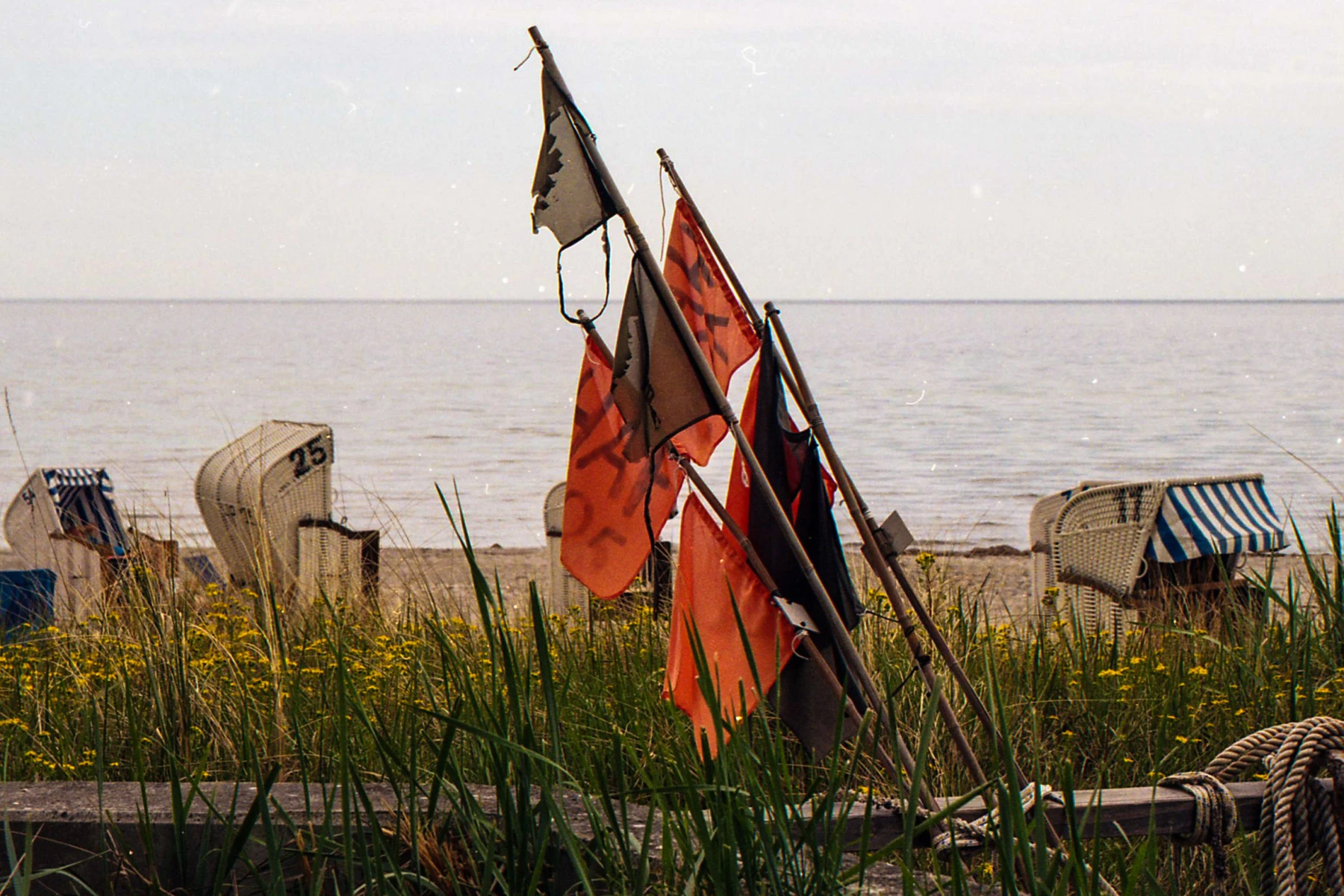 blick aufs meer, im mittelpunkt zerfetzte rote fahnen, die von fischern mal genutzt wurden, jetzt nur noch hier stehen. am strand strandkörbe, im vordergrund dünenpflanzen.