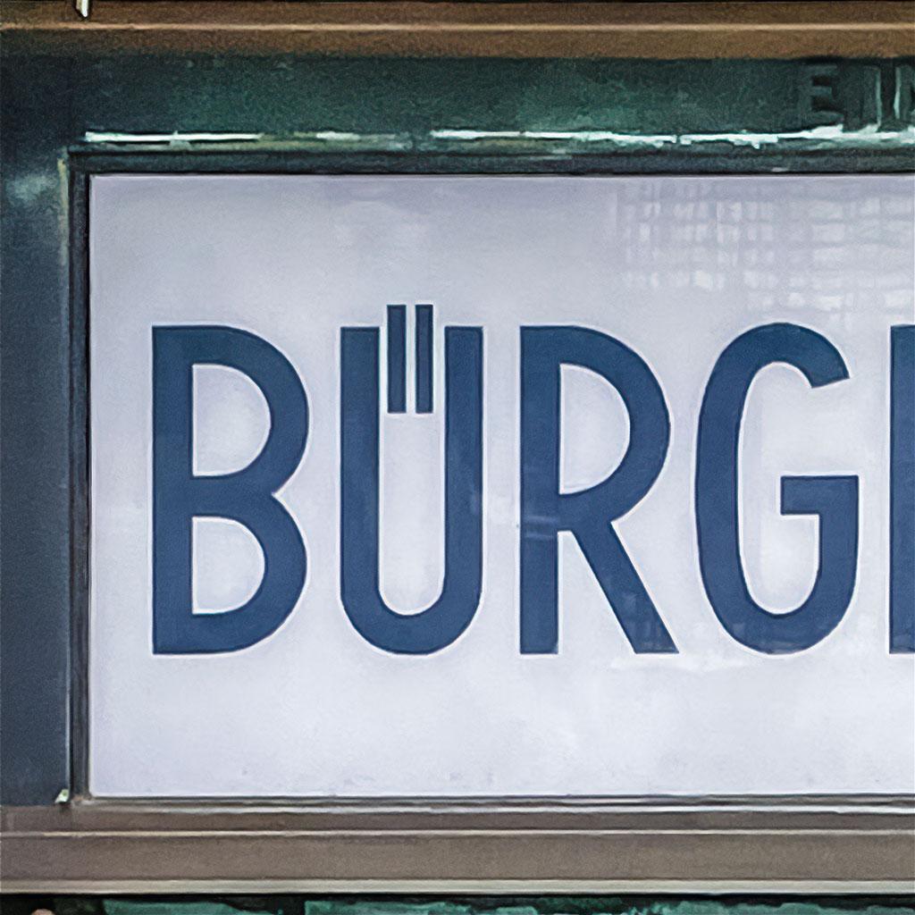Nahaufnahme eines historischen Schildes aus weiß hintermaltem Glas über einem Hauseingang. Im Bildausschnitt die dunkelblauen Großbuchstaben BÜRG mit zwei schmalen senkrechten Strichen als Umlautpunkte, die in die obere Öffnung des U hineinragen