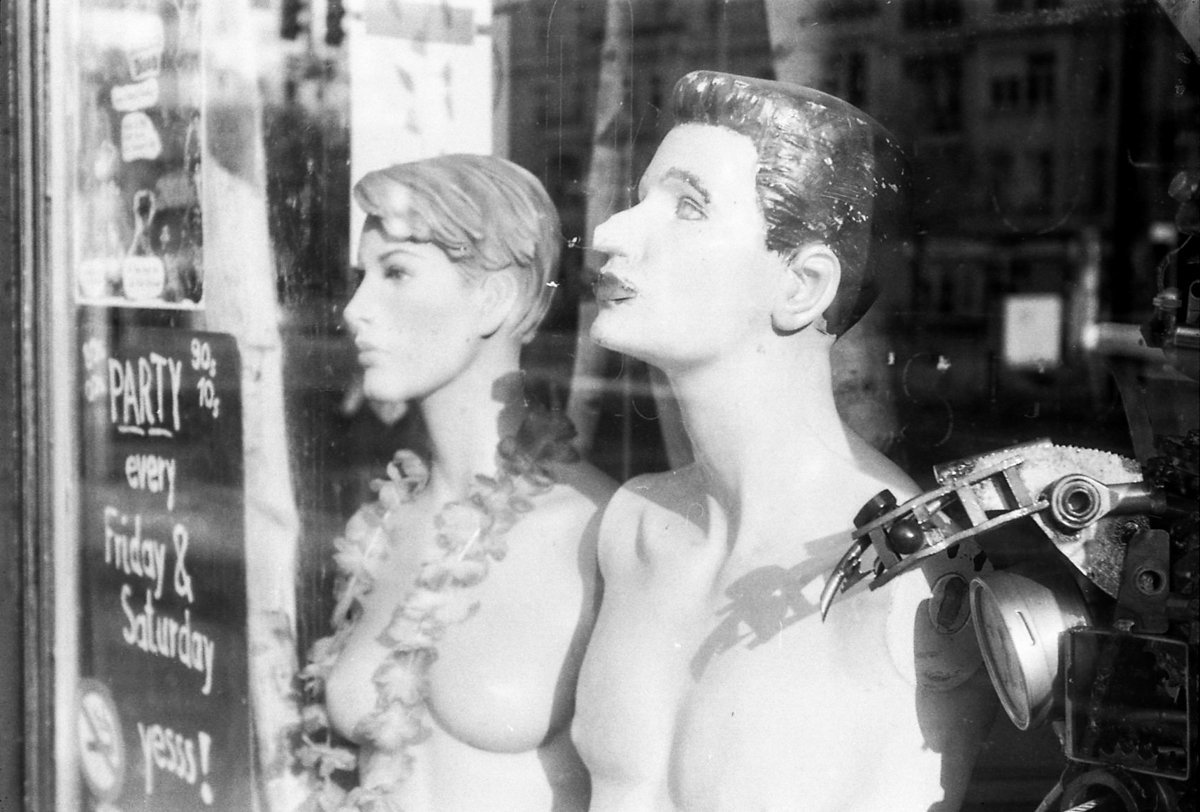 black and white image of two mannequins in shop window looking into sky. sign says "Party every Friday & Saturday yessss!"