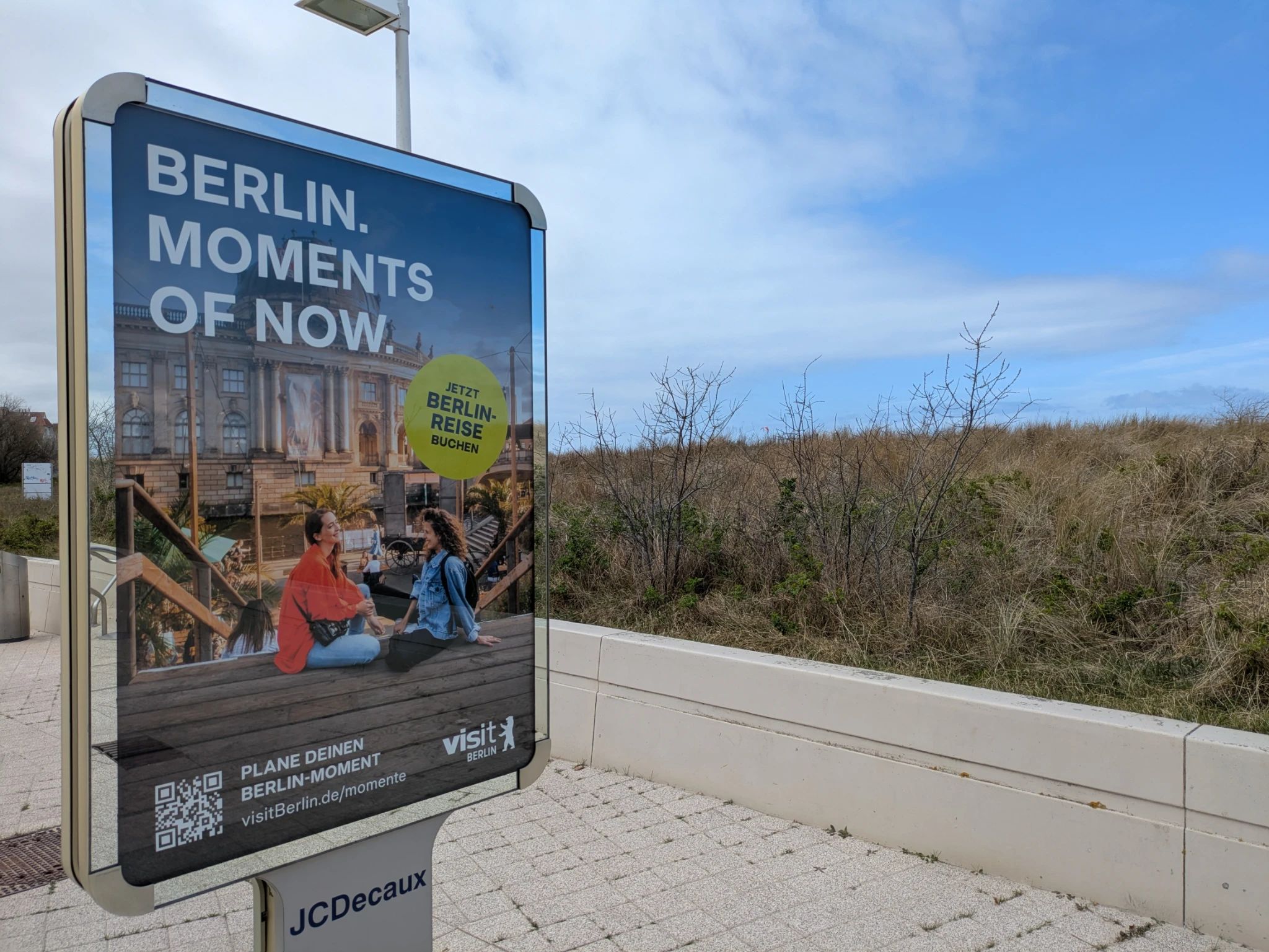 eine plakatwerbung an einer promenade, man sieht düne und blauem himmel. das plakat wirbt 

BERLIN
MOMENTS
OF NOW

und zwei personen sitzen auf einer Treppe vorm Bodemuseum 