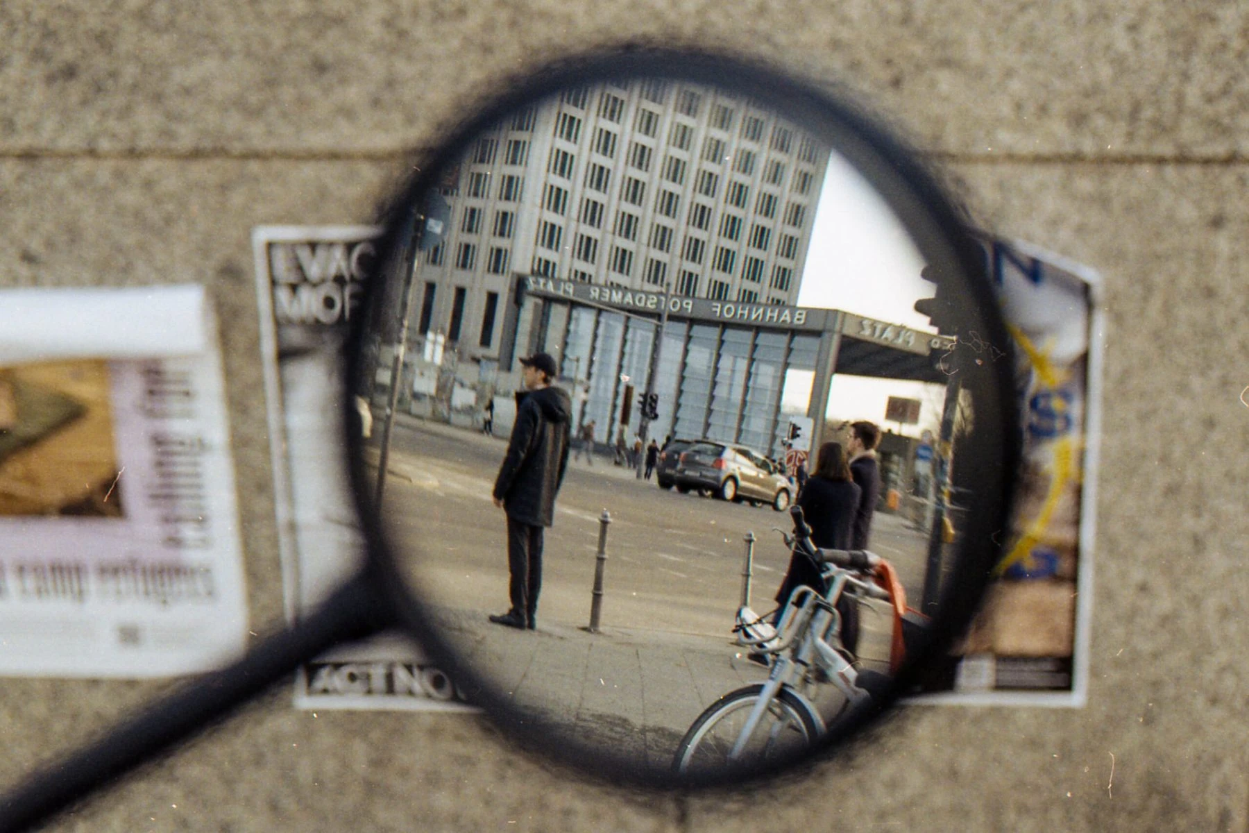 in der bildmitte ein rückspiegel eines fahrrads oder motorrads, es spiegelt sich ein urbaner raum mit modernen gebäuden und menschen. hinter dem spiegel eine plakatwand, unscharf.