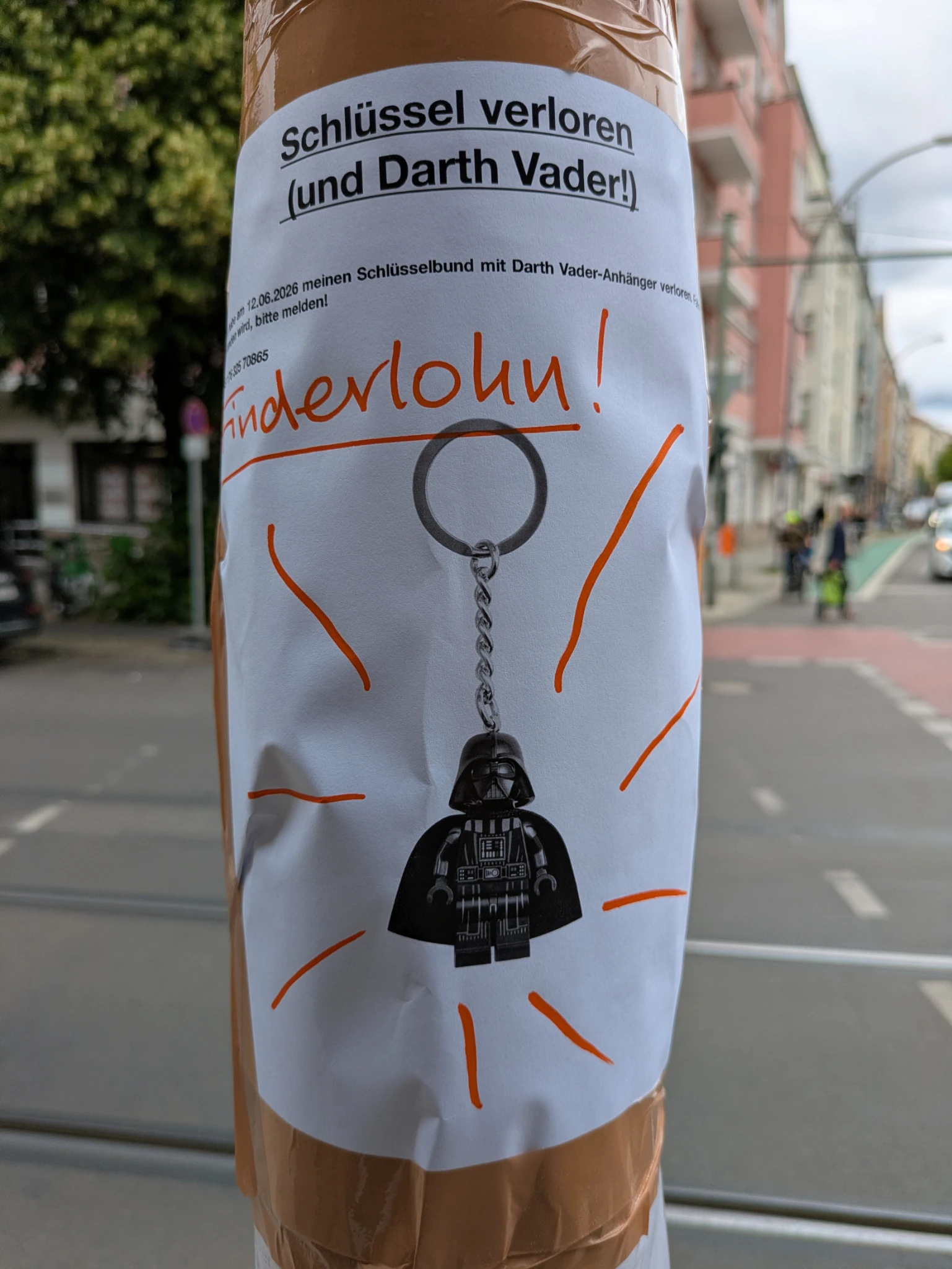 an einer laterne in städtischer umgebung ein zettel, handgeschrieben mit einem bild von einem DARTH VADER-schlüsselanhänger, es wird finderlohn versprochen 