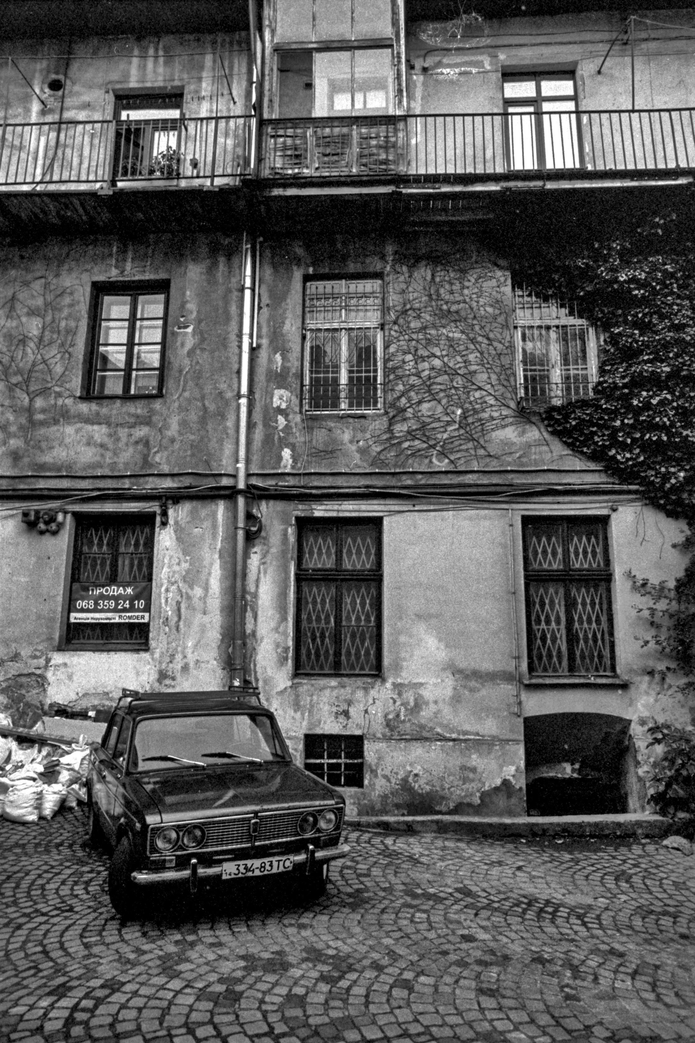 Black-and-white film photograph of a weathered courtyard building facade in Lviv, with peeling plaster, barred windows, a "ПРОДАЖ" (For Sale) sign, wrought-iron balcony railings, bare climbing vines on the wall, and a vintage Lada sedan parked on cobblestones in front.