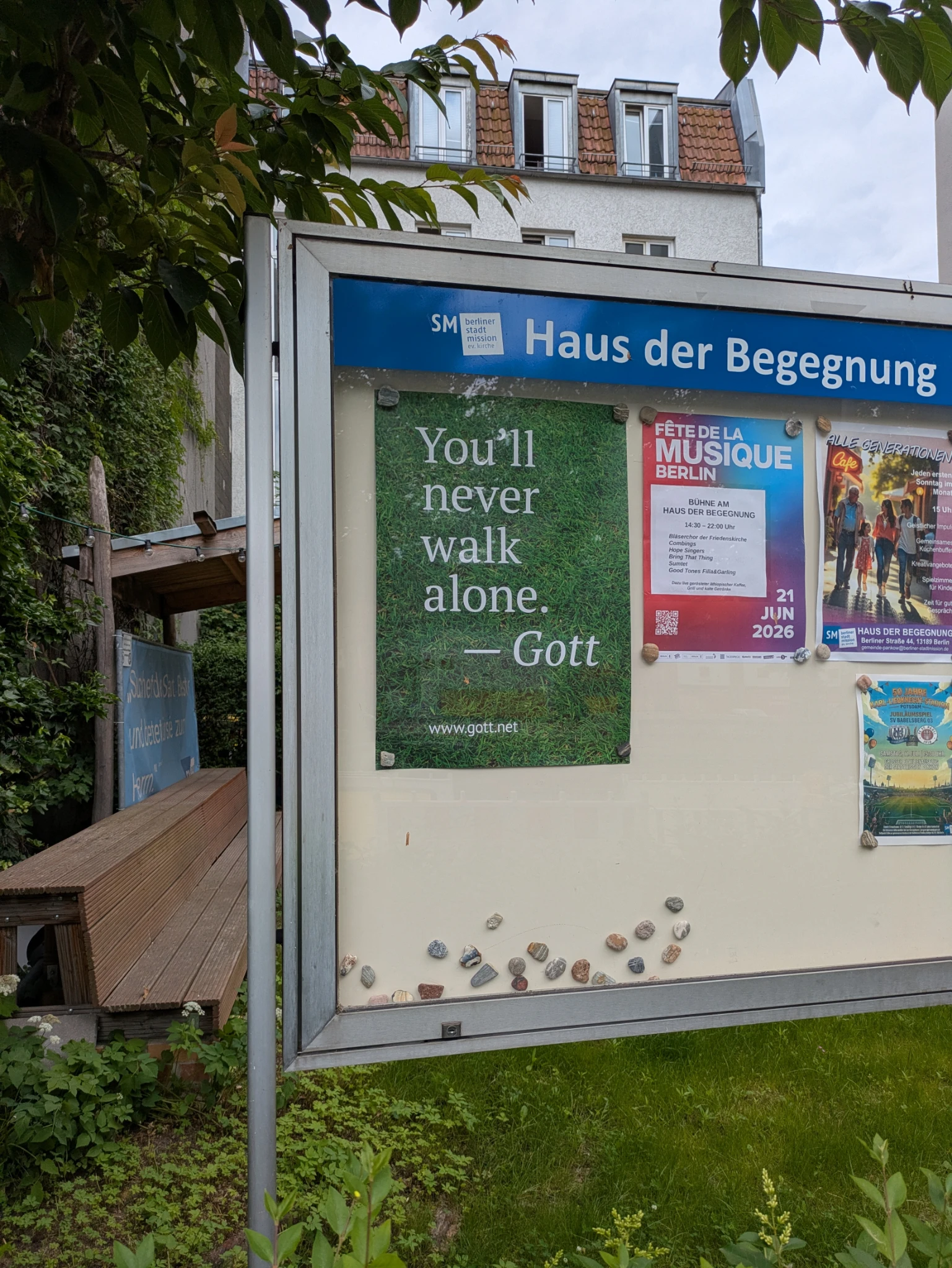 ein infokasten mit infos vor einem gebäude und viel grün drumherum.

HAUS DER BEGEGNUNG, ein grünes plakat:

You'll never walk alone. ~Gott

neben anderen ankündigungen