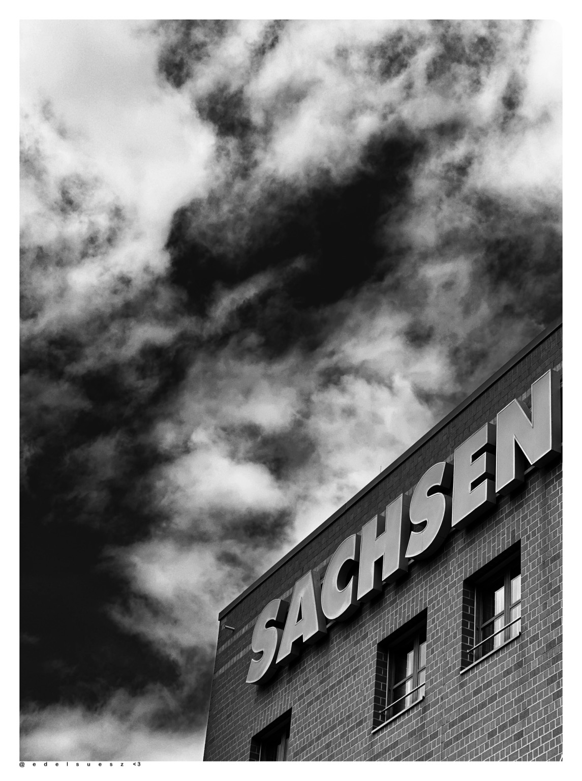 Aufnahme in schwarzweiß, Blick empor an einem großen Gebäude, Flachdach, darunter der Schriftzug in Großbuchstaben „SACHSEN“, darüber dunkler Himmel mit ein paar weißen Wolken 