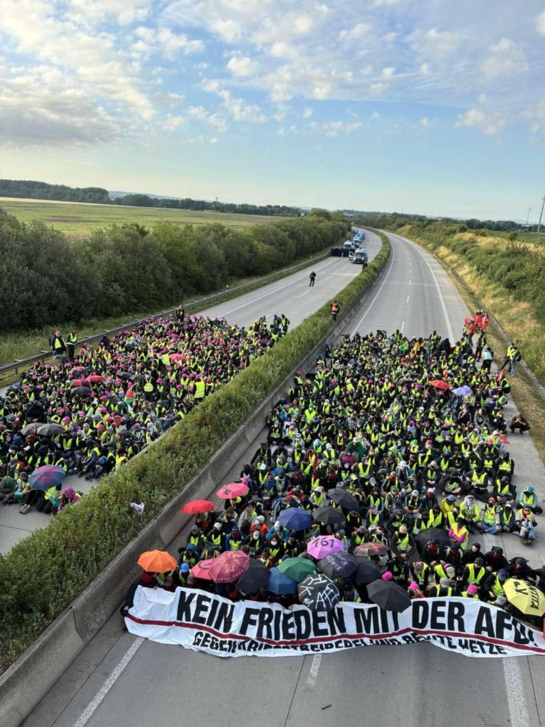 hundreds of anti-fasictactivists are sitting on both sides of the highway. A large banner at the front reads:  Kein Frieden mit der AfD (no peace for AfD). Police vehicles are visible in the distance under a blue, partly cloudy sky
