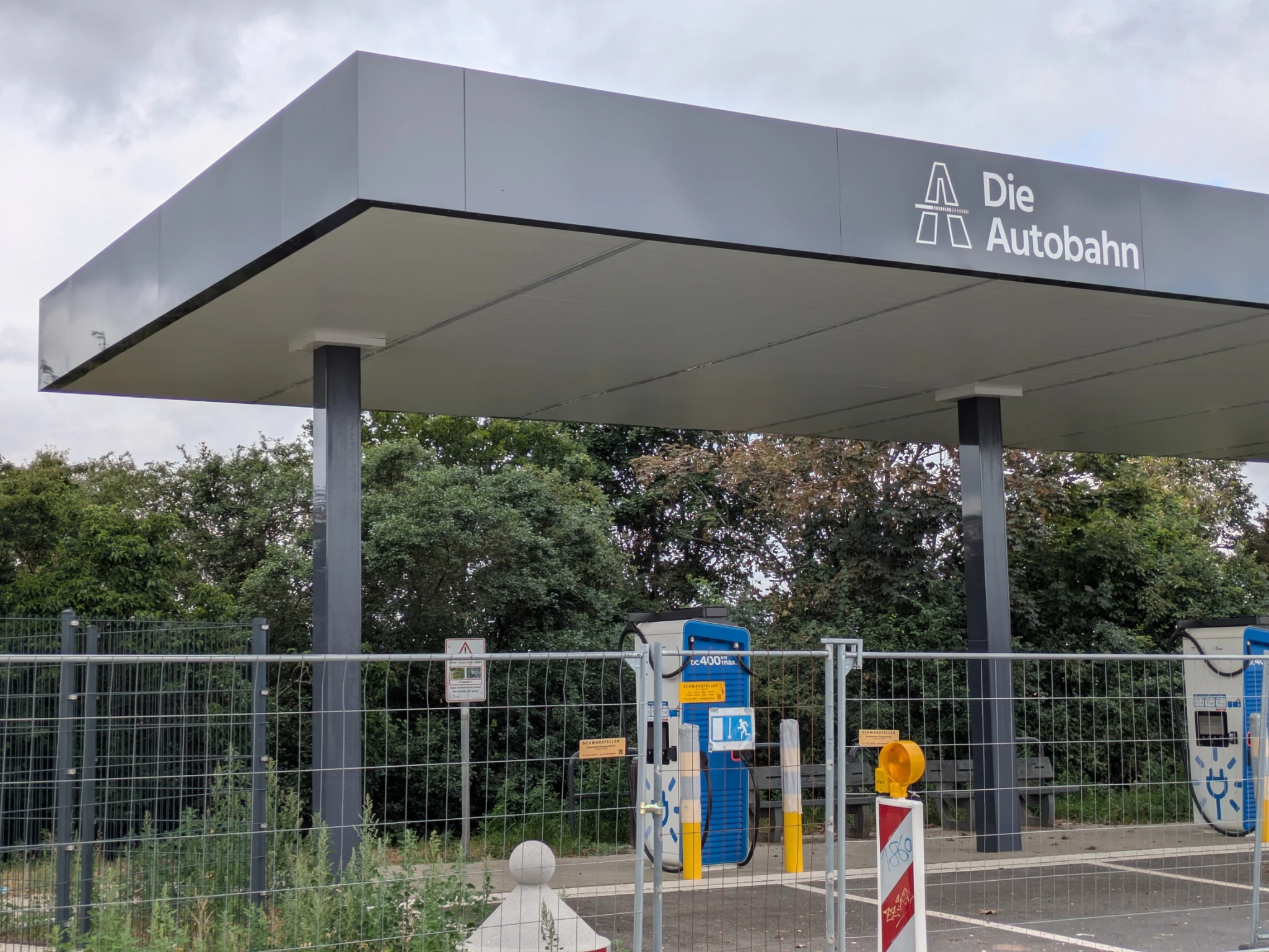 eine überdachte Ladestation für elektrische fahrzeuge, noch im bau inkl bauzaun. am dach steht DIE AUTOBAHN