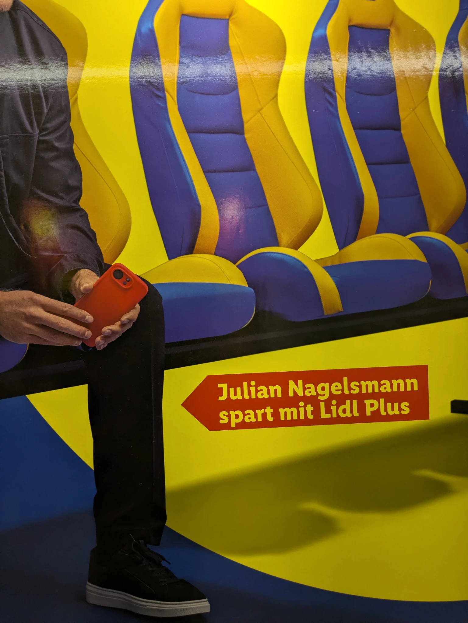 bild eines werbeplakats: typ sitzt auf blau gelben stühlen, man sieht nur bein arm oberkörper, die hand hält ein rotes smartphone. daneben steht 

Julian Nagelsmann spart mit Lidl Plus