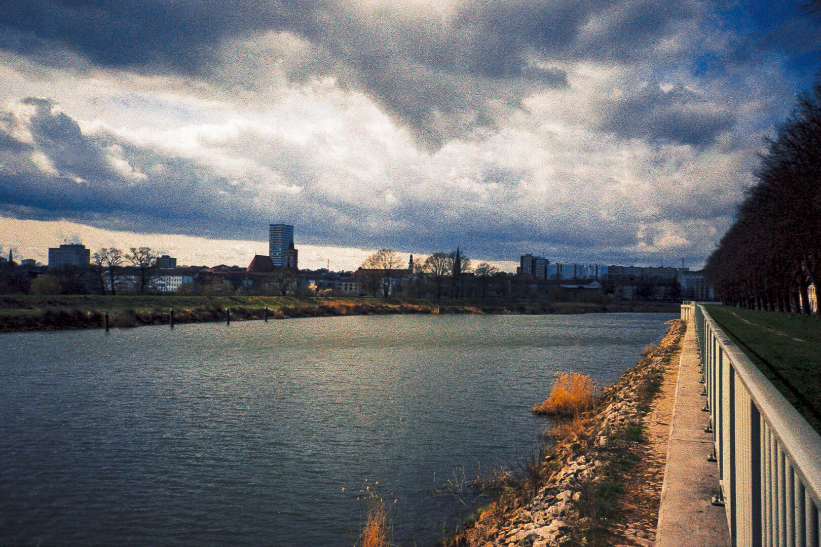 A wide river flows alongside a paved embankment with a white railing, leading towards Frankfurt Oder's skyline under a cloudy sky.