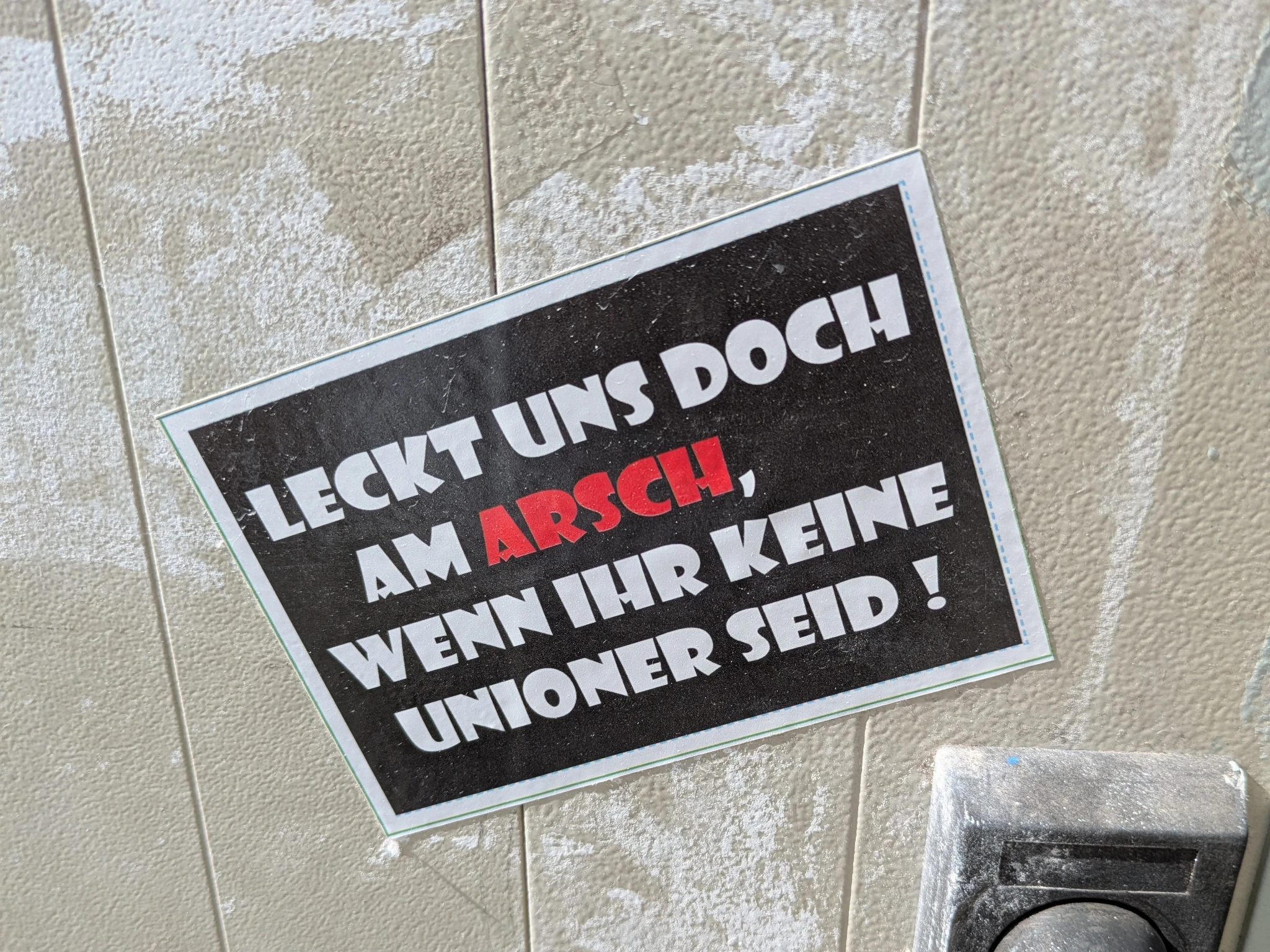 aufkleber auf grauer wand: weiß/rote schrift auf achwarzem grund.

LECKT UNS DOCH AM ARSCH , WENN IHR KEINE UNIONER SEID !