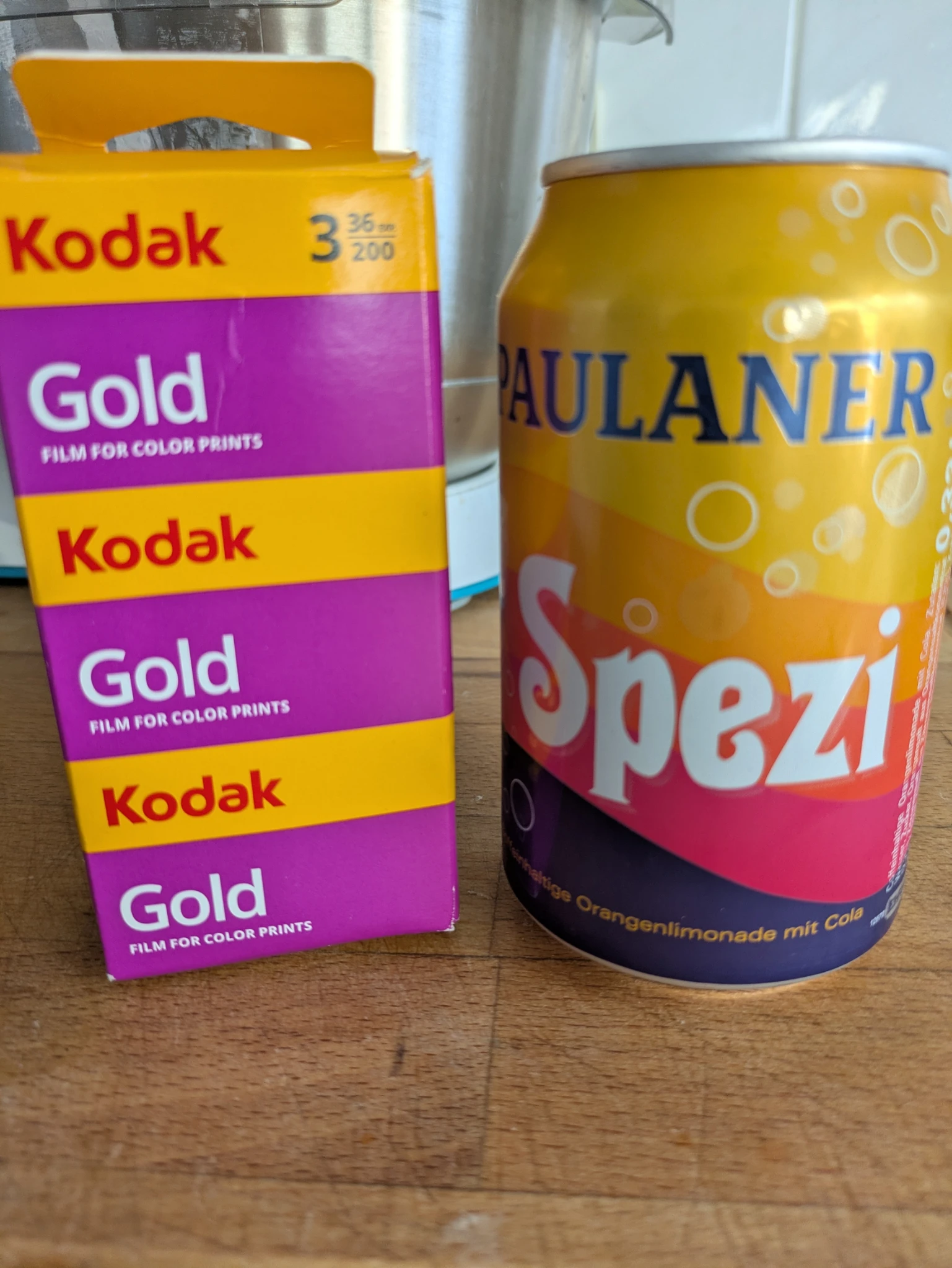 eine Dreierpackung kodak gold in gelb violett, daneben eine dose paulaner spezi in gelb orange rosa dunkelblau