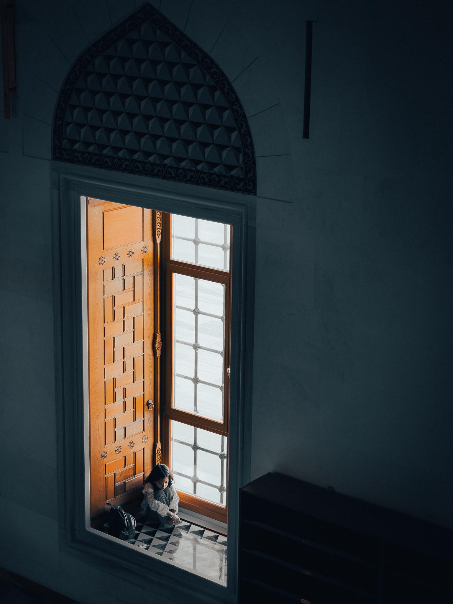 A young girl sits alone in a window, dimly lit despite the bright sun outside. Taken at the Grand Çamlıca Mosque in Istanbul, Türkiye.
